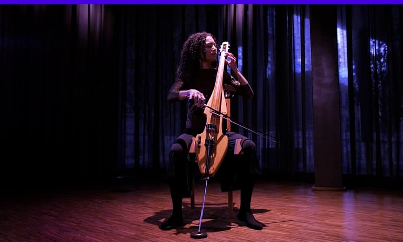 A person sits in a chair on a stage, lit by overhead lighting, and playing a large string instrument.